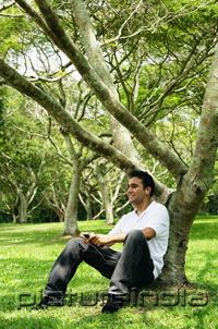 PictureIndia - Young man sitting under tree, listening to music