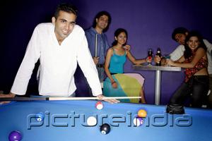 PictureIndia - Young man standing at pool table, people in the background, all looking at camera